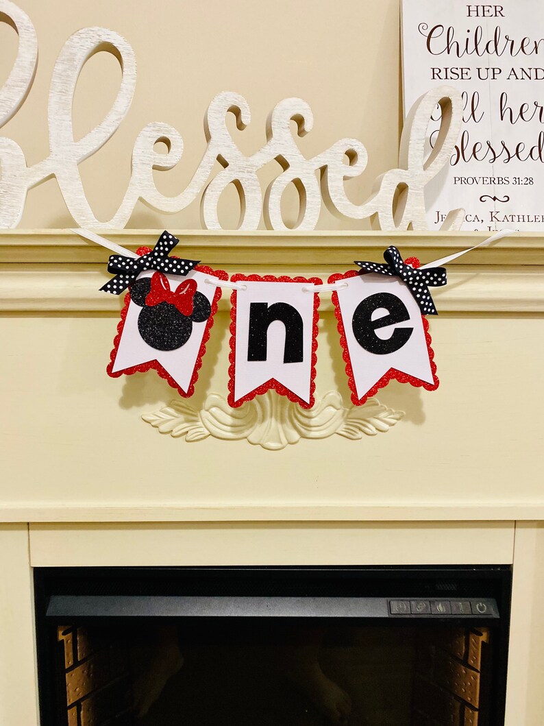 May include: A decorative banner spelling the word one, featuring three white pennants with red scalloped glitter borders. The first pennant displays a black glitter mouse silhouette with a red bow, while the others show black glitter letters.
