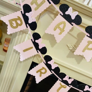 May include: Pink and black birthday banner with gold glitter letters spelling "Happy Birthday". Each letter is attached to a black Mickey Mouse ear with a pink bow.