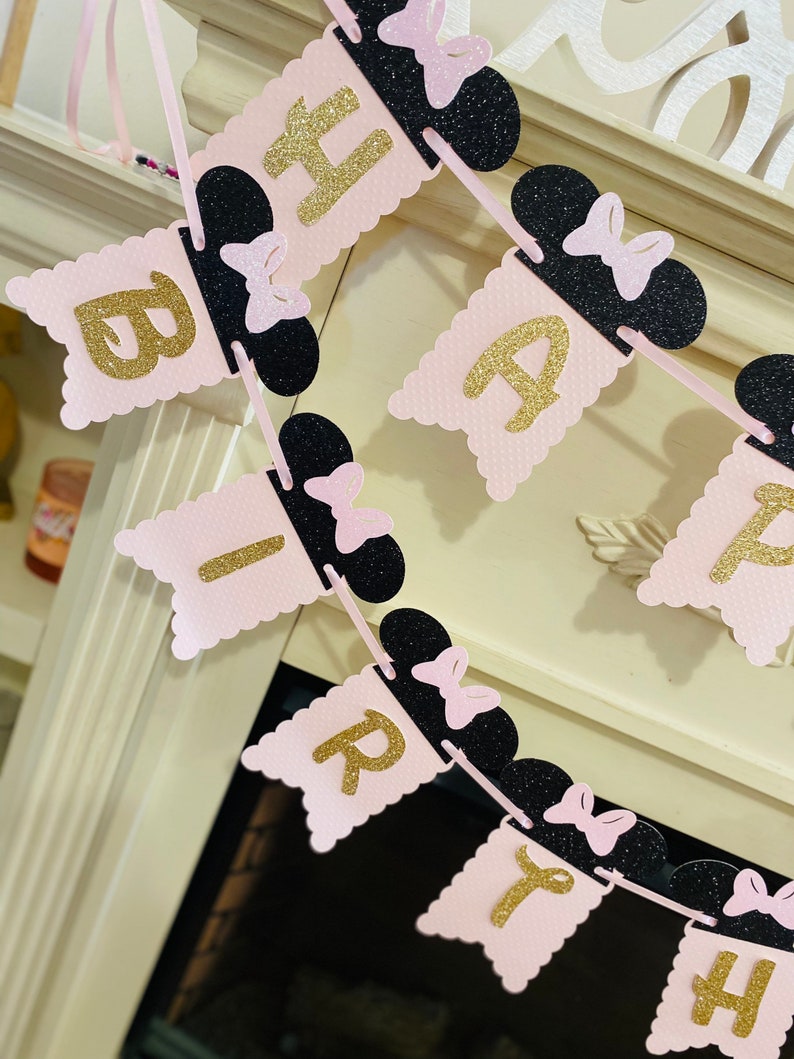 May include: Pink and black birthday banner with gold glitter letters spelling "Happy Birthday". Each letter is attached to a black Mickey Mouse ear with a pink bow.