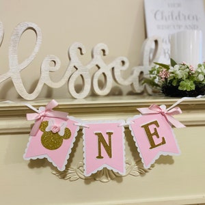 Puede incluir: Pancarta de "Uno" con purpurina rosa y dorada con una cabeza de Minnie Mouse en la primera bandera. La pancarta est&aacute; colgada en una repisa blanca con un letrero de madera que dice "Bendito" y un jarr&oacute;n blanco con vegetaci&oacute;n.