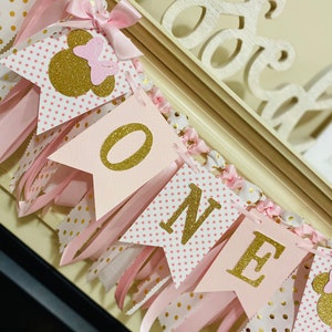 May include: A pink and gold "ONE" banner with Minnie Mouse accents. The banner features pink pennants with gold glitter lettering and polka dots, accented with pink ribbons and bows. The banner is designed for a first birthday celebration.