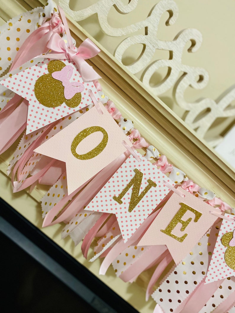 May include: A pink and gold "ONE" banner with Minnie Mouse accents. The banner features pink pennants with gold glitter lettering and polka dots, accented with pink ribbons and bows. The banner is designed for a first birthday celebration.