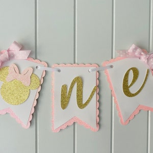 Puede incluir: Pancarta de "uno" con purpurina rosa y dorada con una cabeza de Minnie Mouse de purpurina rosa y dorada en el lado izquierdo. La pancarta est&aacute; enhebrada con cinta blanca y lazos de lunares rosa y blancos.