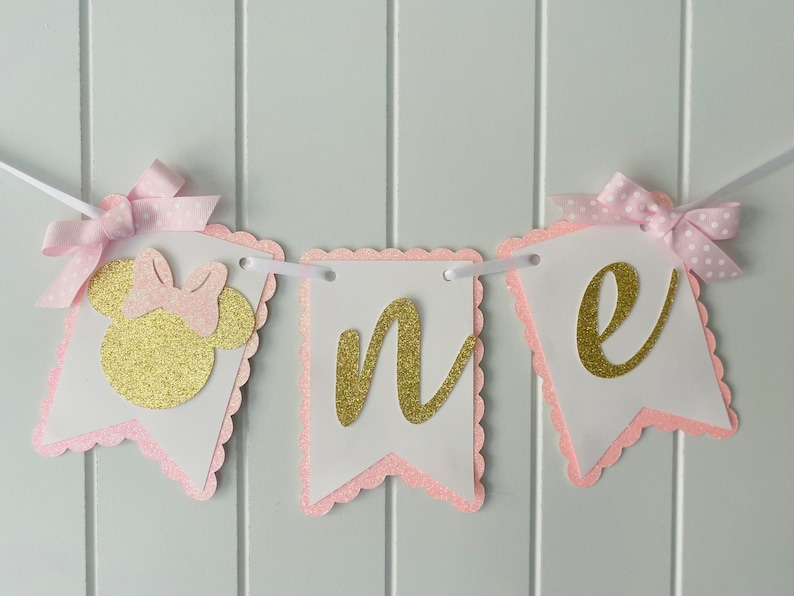 Puede incluir: Pancarta de "uno" con purpurina rosa y dorada con una cabeza de Minnie Mouse de purpurina rosa y dorada en el lado izquierdo. La pancarta est&aacute; enhebrada con cinta blanca y lazos de lunares rosa y blancos.