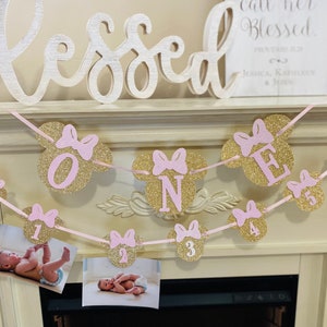 May include: A pink and gold glitter banner with Mickey Mouse ears and numbers from 0 to 6. The banner says "Blessed" and has a pink ribbon.