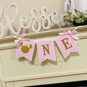 Puede incluir: Pancarta de "UNO" de color rosa y dorado con purpurina y borde blanco festoneado. La pancarta presenta una cabeza de Minnie Mouse rosa con un lazo de purpurina dorada y una flor rosa.