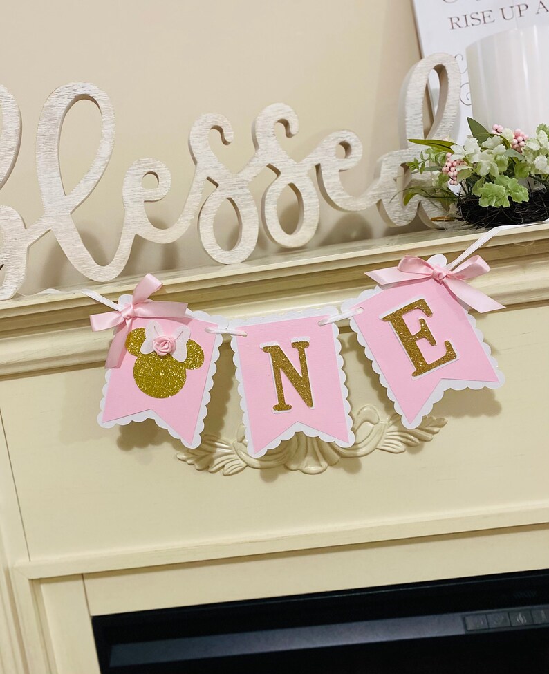 Puede incluir: Pancarta de "UNO" de color rosa y dorado con purpurina y borde blanco festoneado. La pancarta presenta una cabeza de Minnie Mouse rosa con un lazo de purpurina dorada y una flor rosa.