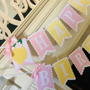 May include: A pink and yellow banner with the words "Happy Birthday" in white letters. The banner has a scalloped edge and is decorated with small pink bows and a lemon illustration.
