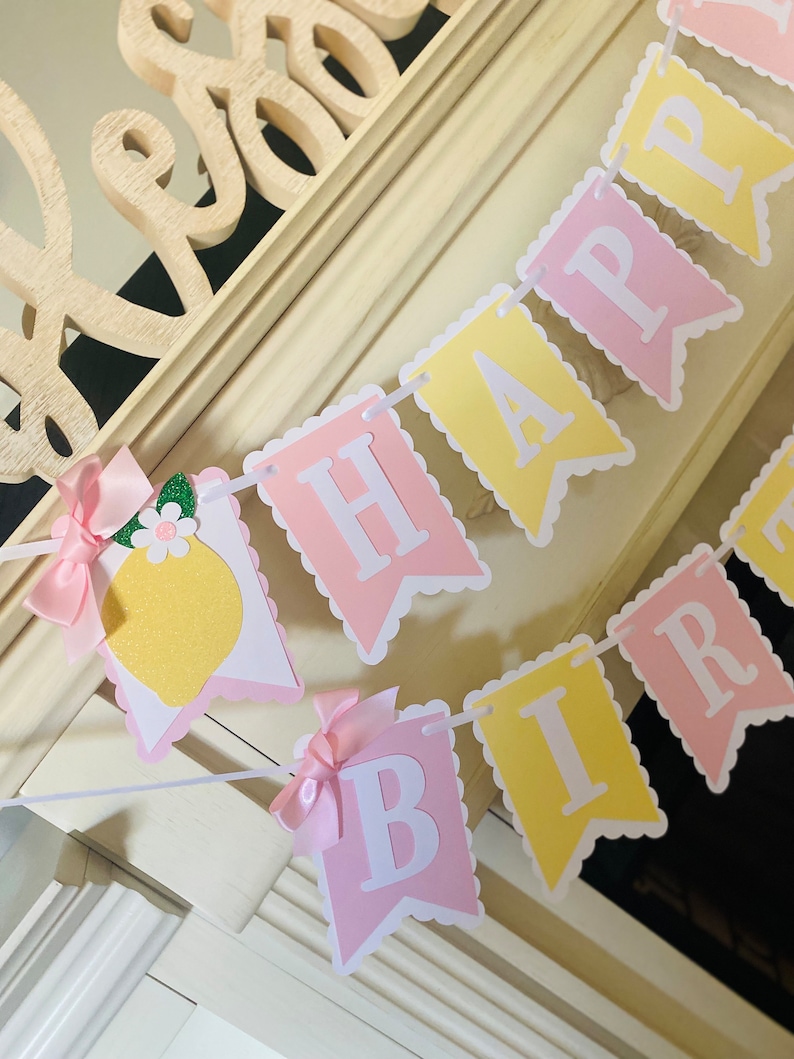 May include: A pink and yellow banner with the words "Happy Birthday" in white letters. The banner has a scalloped edge and is decorated with small pink bows and a lemon illustration.