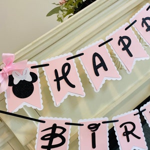 Puede incluir: Una pancarta rosa y negra con bordes festoneados que dice "Happy Birthday". La pancarta est&aacute; enhebrada con una cuerda negra y presenta una cabeza de Minnie Mouse negra y blanca con un lazo rosa.