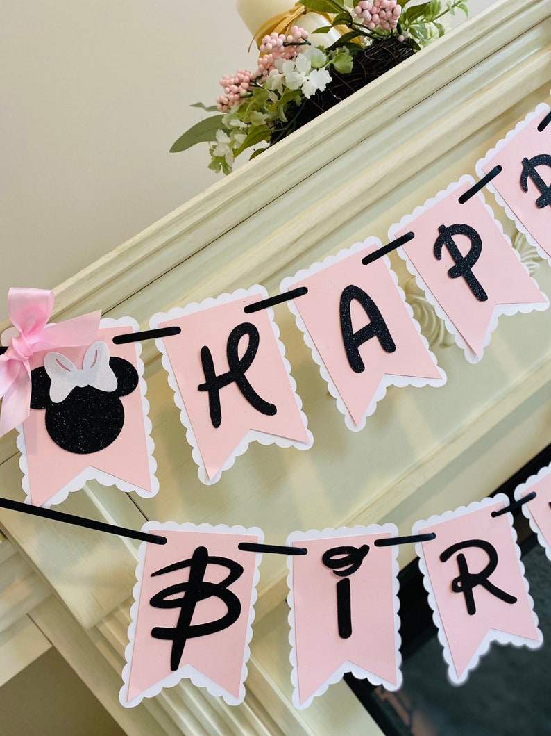 Puede incluir: Una pancarta rosa y negra con bordes festoneados que dice "Happy Birthday". La pancarta est&aacute; enhebrada con una cuerda negra y presenta una cabeza de Minnie Mouse negra y blanca con un lazo rosa.