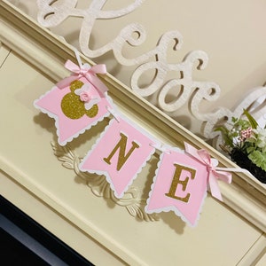 Puede incluir: Bandera rosa y dorada con la palabra "ONE" en letras doradas con purpurina. La bandera est&aacute; decorada con un lazo rosa y una cabeza de Mickey Mouse dorada con purpurina.