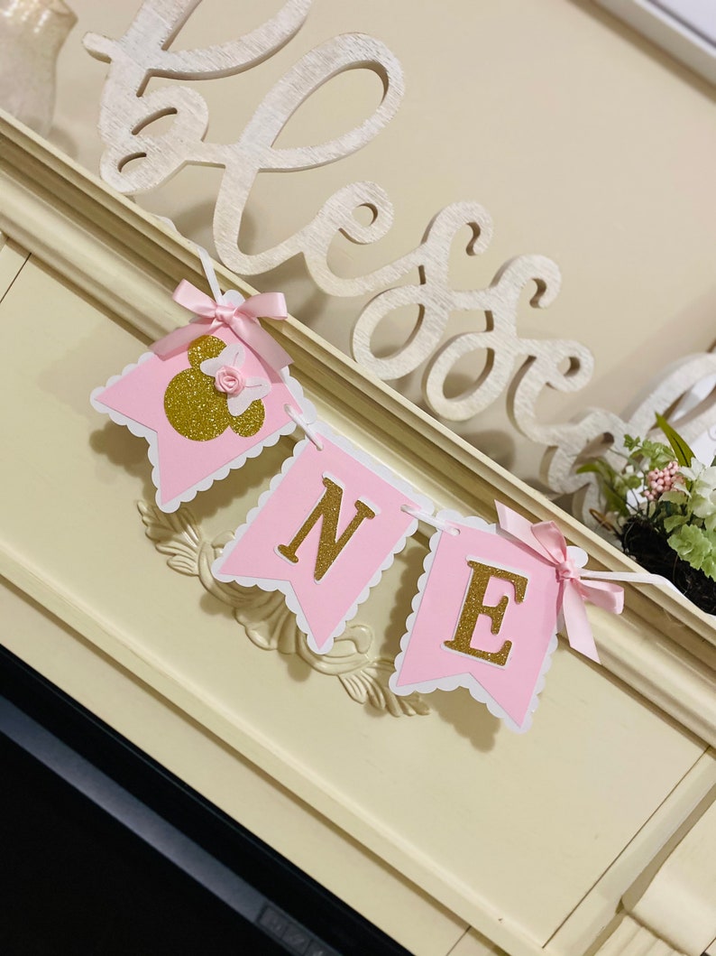 Puede incluir: Bandera rosa y dorada con la palabra "ONE" en letras doradas con purpurina. La bandera est&aacute; decorada con un lazo rosa y una cabeza de Mickey Mouse dorada con purpurina.