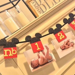 May include: A Mickey Mouse themed baby milestone banner with black glitter ears and red and yellow numbers. The banner includes photos of a baby at different ages. The banner reads "nb, 1, 2".