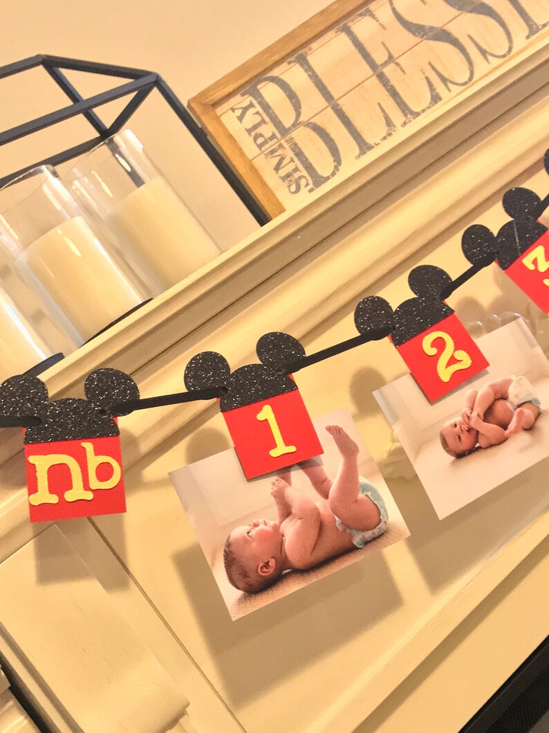 May include: A Mickey Mouse themed baby milestone banner with black glitter ears and red and yellow numbers. The banner includes photos of a baby at different ages. The banner reads "nb, 1, 2".
