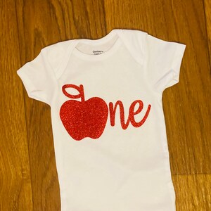 May include: White onesie with a red glitter apple and the word "one" in red glitter.