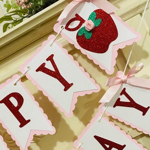 May include: A pink and white banner with red glitter letters spelling "Happy Day" with a red glitter apple with a green leaf and a pink rose on top. The banner has pink scalloped edges and pink bows.