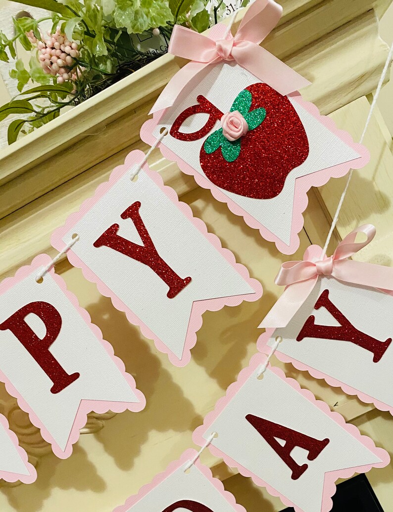 May include: A pink and white banner with red glitter letters spelling "Happy Day" with a red glitter apple with a green leaf and a pink rose on top. The banner has pink scalloped edges and pink bows.