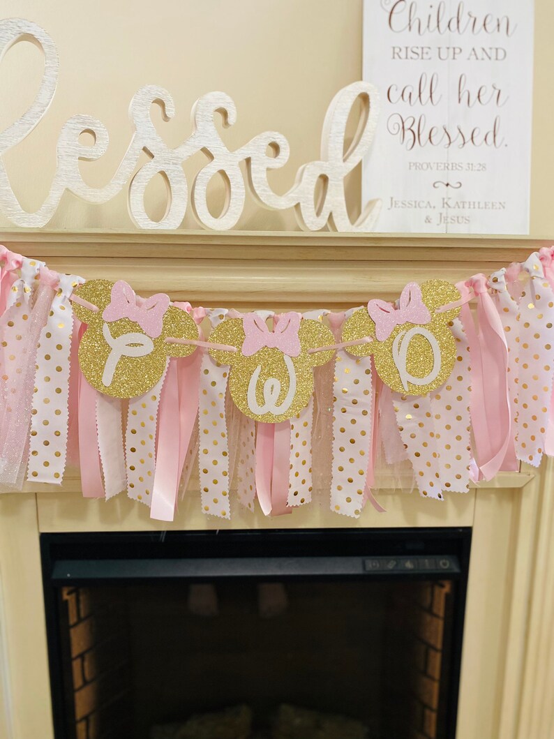 May include: A decorative banner with gold glitter Mickey Mouse head cutouts spelling "TWO". The banner is adorned with pink bows and ribbons, and is set against a backdrop of pink and gold polka dot fabric. A sign reads "Children rise up and call her Blessed."