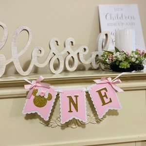 Puede incluir: Pancarta rosa y dorada "Uno" con una cabeza de Minnie Mouse en la primera bandera. La pancarta est&aacute; colgada en una repisa blanca con un letrero de madera blanca que dice "Bendecido" y una vela blanca en un tarro de vidrio.