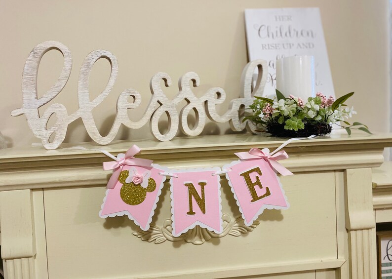 Puede incluir: Pancarta rosa y dorada "Uno" con una cabeza de Minnie Mouse en la primera bandera. La pancarta est&aacute; colgada en una repisa blanca con un letrero de madera blanca que dice "Bendecido" y una vela blanca en un tarro de vidrio.