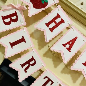 May include: A red and white banner with the words "Happy Birthday" in red glitter letters. The banner is decorated with pink scalloped edges and a red apple with a green leaf and a pink flower.