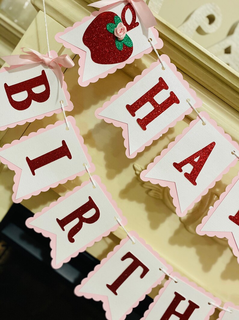May include: A red and white banner with the words "Happy Birthday" in red glitter letters. The banner is decorated with pink scalloped edges and a red apple with a green leaf and a pink flower.