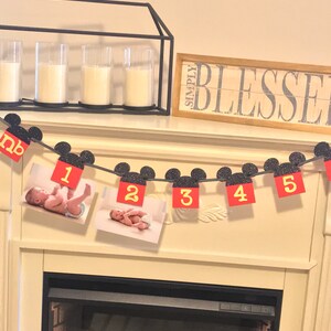 May include: A red and black Mickey Mouse themed banner with photos of a baby and the numbers 1 through 5. The banner is hanging above a fireplace.