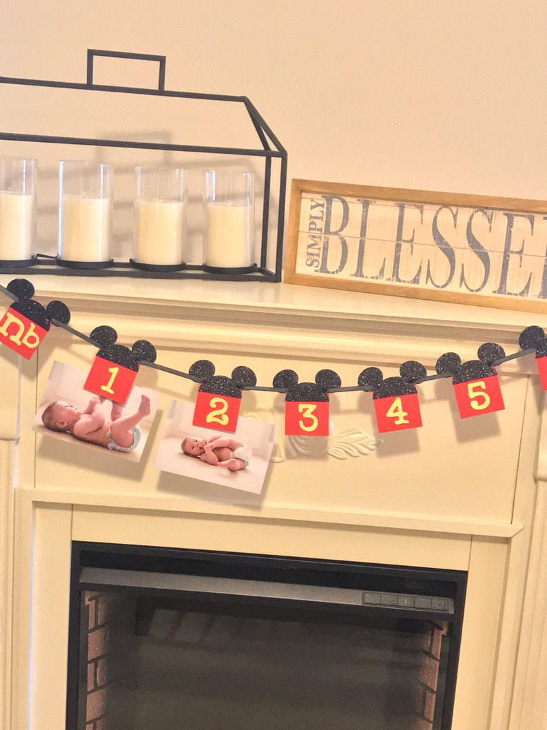May include: A red and black Mickey Mouse themed banner with photos of a baby and the numbers 1 through 5. The banner is hanging above a fireplace.
