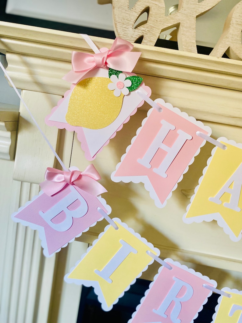 May include: A pink and yellow birthday banner with the word "Happy Birthday" in white letters. The banner has a scalloped edge and is decorated with a lemon and a bow.