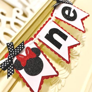 May include: A decorative banner spelling the word ONE, featuring white pennants with red glitter scalloped edges. The letter O is replaced by a black glitter mouse silhouette with a red glitter bow, accented by a black and white polka dot ribbon.