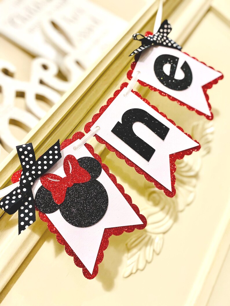 May include: A decorative banner spelling the word ONE, featuring white pennants with red glitter scalloped edges. The letter O is replaced by a black glitter mouse silhouette with a red glitter bow, accented by a black and white polka dot ribbon.