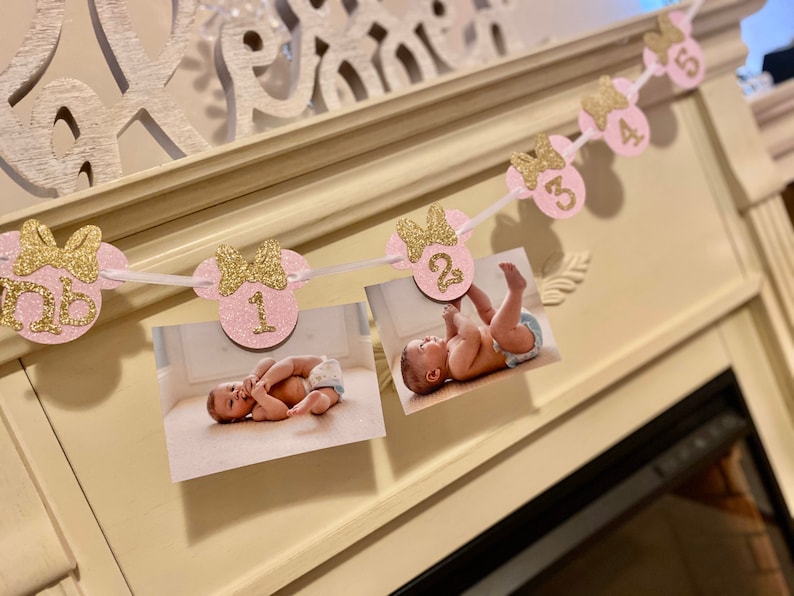 May include: Pink and gold glitter Minnie Mouse themed photo banner with numbers 1 through 5. Each number is on a separate pink circle with a gold bow. The banner is strung with white ribbon and has photos of a baby girl at each number.
