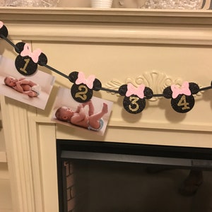 May include: A black and gold glitter banner with Minnie Mouse head cutouts, pink bows, and numbers 1-5. The banner includes photos of a baby. The banner is strung across a fireplace.