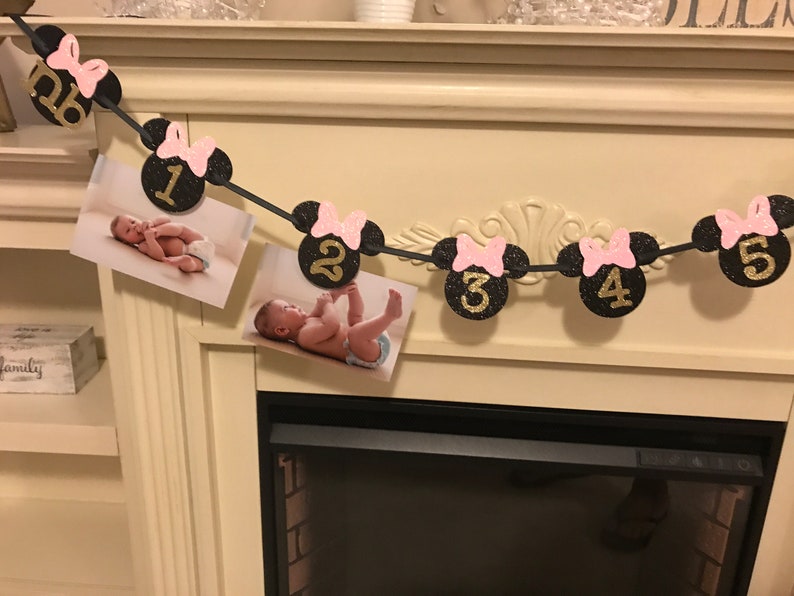 May include: A black and gold glitter banner with Minnie Mouse head cutouts, pink bows, and numbers 1-5. The banner includes photos of a baby. The banner is strung across a fireplace.