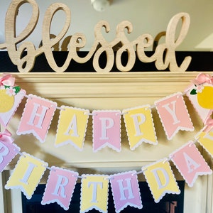 May include: A pink and yellow banner with the words "Happy Birthday" strung across a white mantel. The banner is decorated with lemons and bows. The word "Blessed" is written on a wooden sign above the banner.
