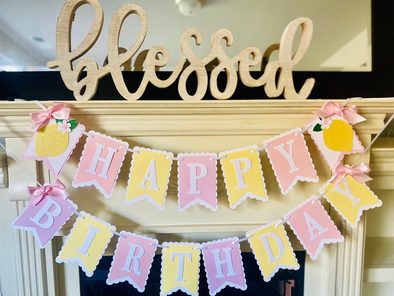 May include: A pink and yellow banner with the words "Happy Birthday" strung across a white mantel. The banner is decorated with lemons and bows. The word "Blessed" is written on a wooden sign above the banner.