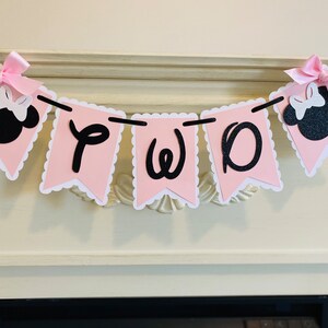 May include: Pink and black banner with the word "TWO" and Minnie Mouse heads on each end. The banner is strung on a white fireplace mantel.