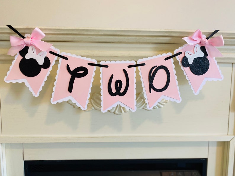 May include: Pink and black banner with the word "TWO" and Minnie Mouse heads on each end. The banner is strung on a white fireplace mantel.