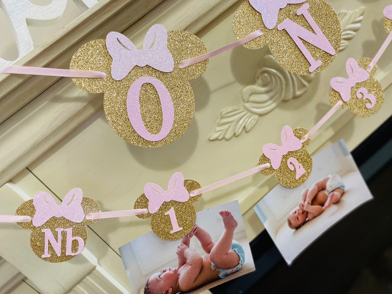 May include: A pink and gold glitter banner with a Minnie Mouse theme. The banner has the letters "Nb", "1", "2", "3", and "One" on it. The banner is decorated with pink bows.