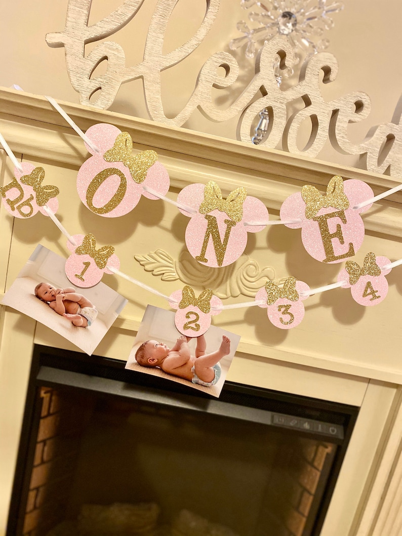 Op de afbeelding: Een roze en gouden glitterbanner met Minnie Mouse-thema met het woord "ONE" en de nummers 1-4. De banner is aan een wit lint geregen en heeft in elke sectie foto's van een baby.