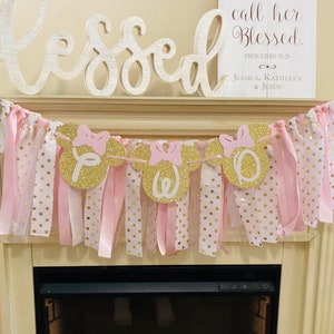 May include: A decorative banner with gold glitter Minnie Mouse head cutouts spelling "TWO" with pink bows. The banner is strung with pink and white polka dot fabric and ribbons. A wooden sign reads "Her Children Rise Up and call her Blessed."