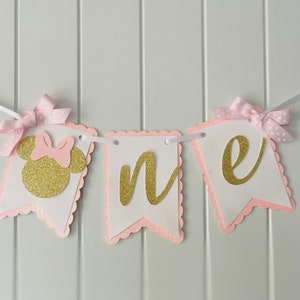 Puede incluir: Pancarta de "uno" con purpurina rosa y dorada con una cabeza de Minnie Mouse de purpurina rosa y dorada. La pancarta es perfecta para una fiesta de primer cumplea&ntilde;os.