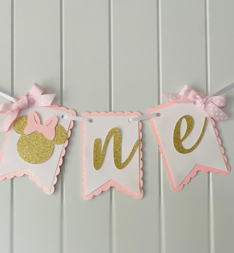 Puede incluir: Pancarta de "uno" con purpurina rosa y dorada con una cabeza de Minnie Mouse de purpurina rosa y dorada. La pancarta es perfecta para una fiesta de primer cumplea&ntilde;os.