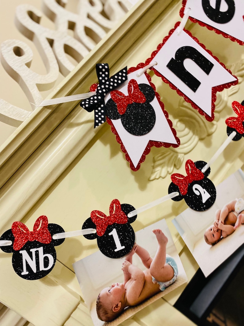 May include: A red and black Minnie Mouse themed banner with the word "one" and numbers 1 and 2. The banner is decorated with red bows and black polka dots. There are photos of a baby in the banner.