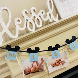 Pode incluir: Uma faixa de Mickey Mouse em azul e preto com n&uacute;meros dourados brilhantes 1, 2 e 3. A faixa est&aacute; pendurada em uma lareira branca com a palavra "aben&ccedil;oado" em letras brancas na lareira.