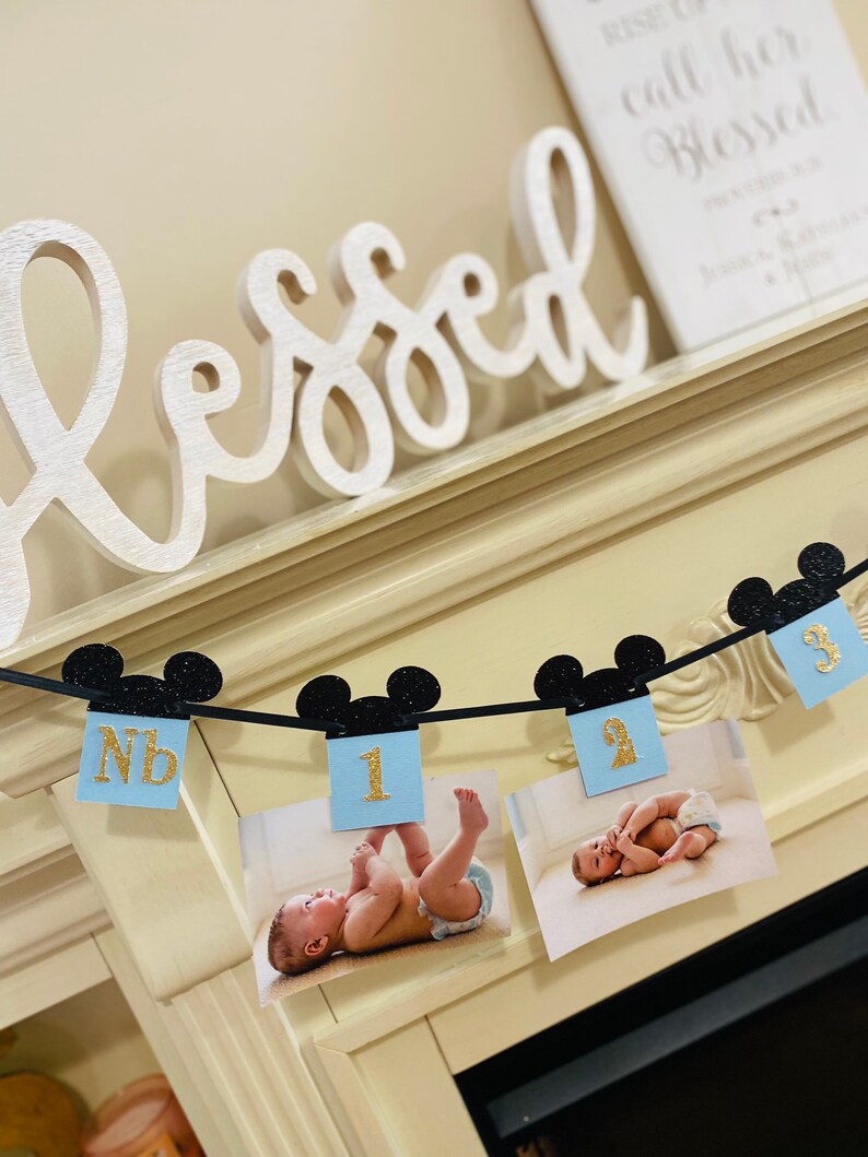 Pode incluir: Uma faixa de Mickey Mouse em azul e preto com n&uacute;meros dourados brilhantes 1, 2 e 3. A faixa est&aacute; pendurada em uma lareira branca com a palavra "aben&ccedil;oado" em letras brancas na lareira.