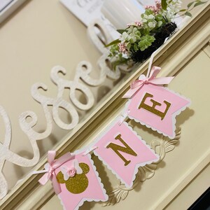 Puede incluir: Una pancarta rosa y dorada con la palabra "ONE" en letras doradas y una cabeza de Minnie Mouse rosa y dorada en el lado izquierdo. La pancarta est&aacute; colgada en una superficie blanca.