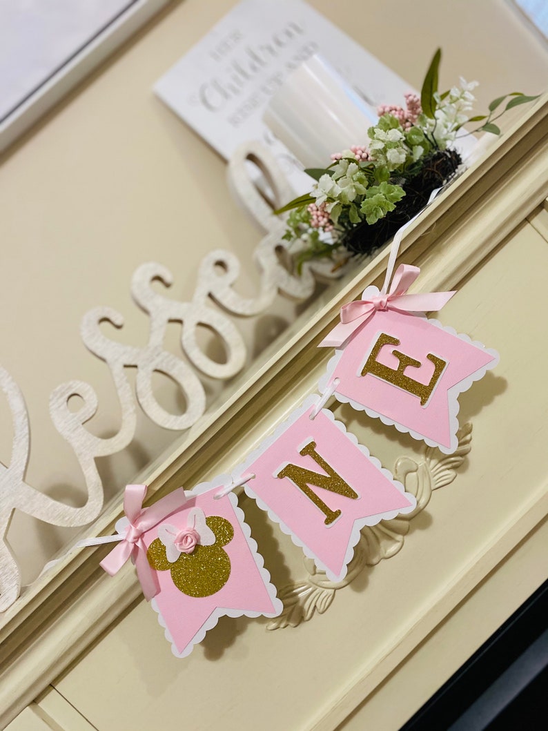 Puede incluir: Una pancarta rosa y dorada con la palabra "ONE" en letras doradas y una cabeza de Minnie Mouse rosa y dorada en el lado izquierdo. La pancarta est&aacute; colgada en una superficie blanca.