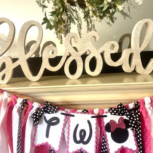 May include: A decorative banner with pink, white, and black polka dot and striped fabric strips. The banner includes three white pennants with the letters "P", "W", and a Minnie Mouse silhouette. A wooden "Blessed" sign and a floral wreath are in the background.